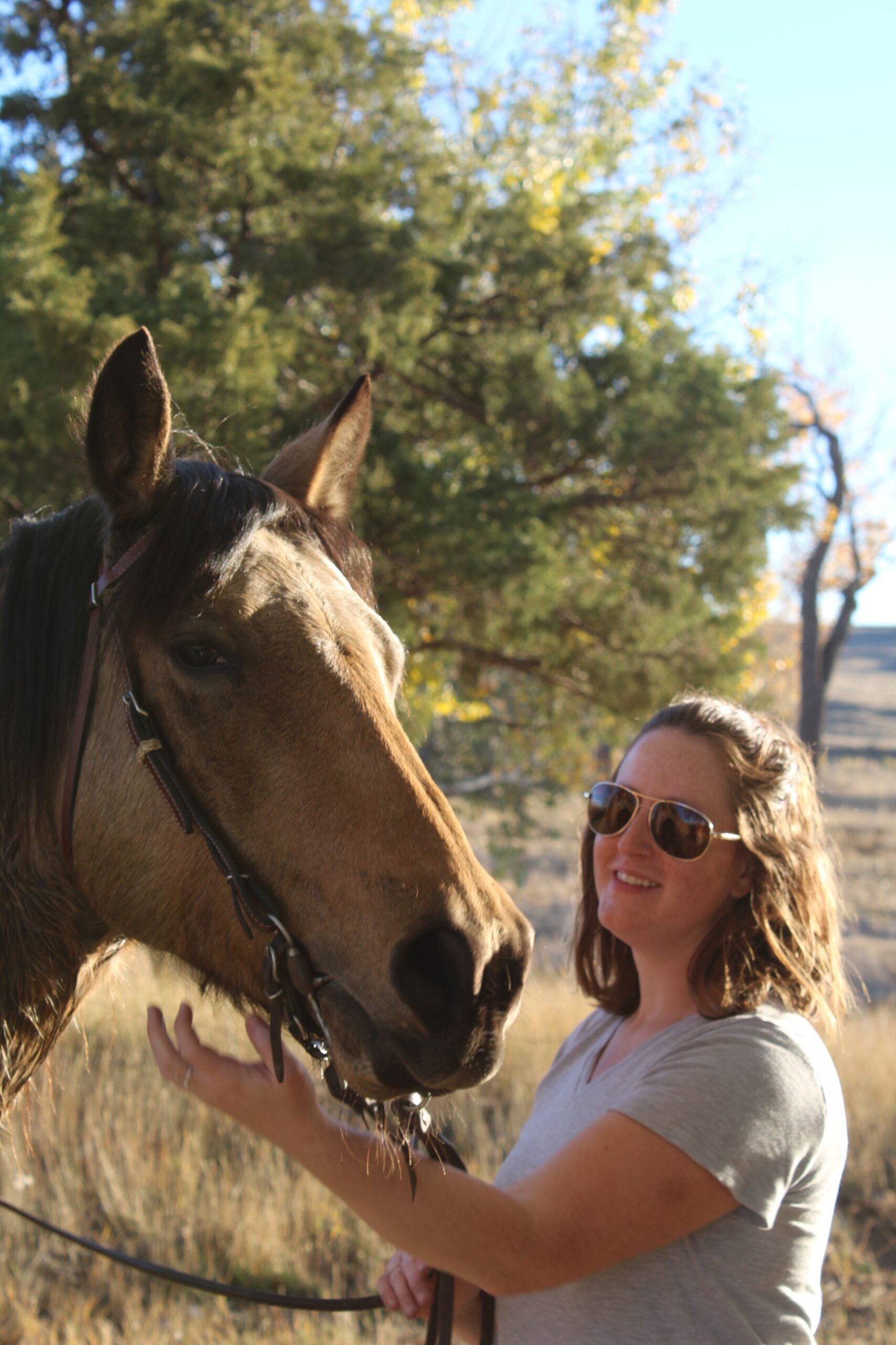 A young woman in sunglasses poses with a buckskin Morgan mare on a fall day in the country.